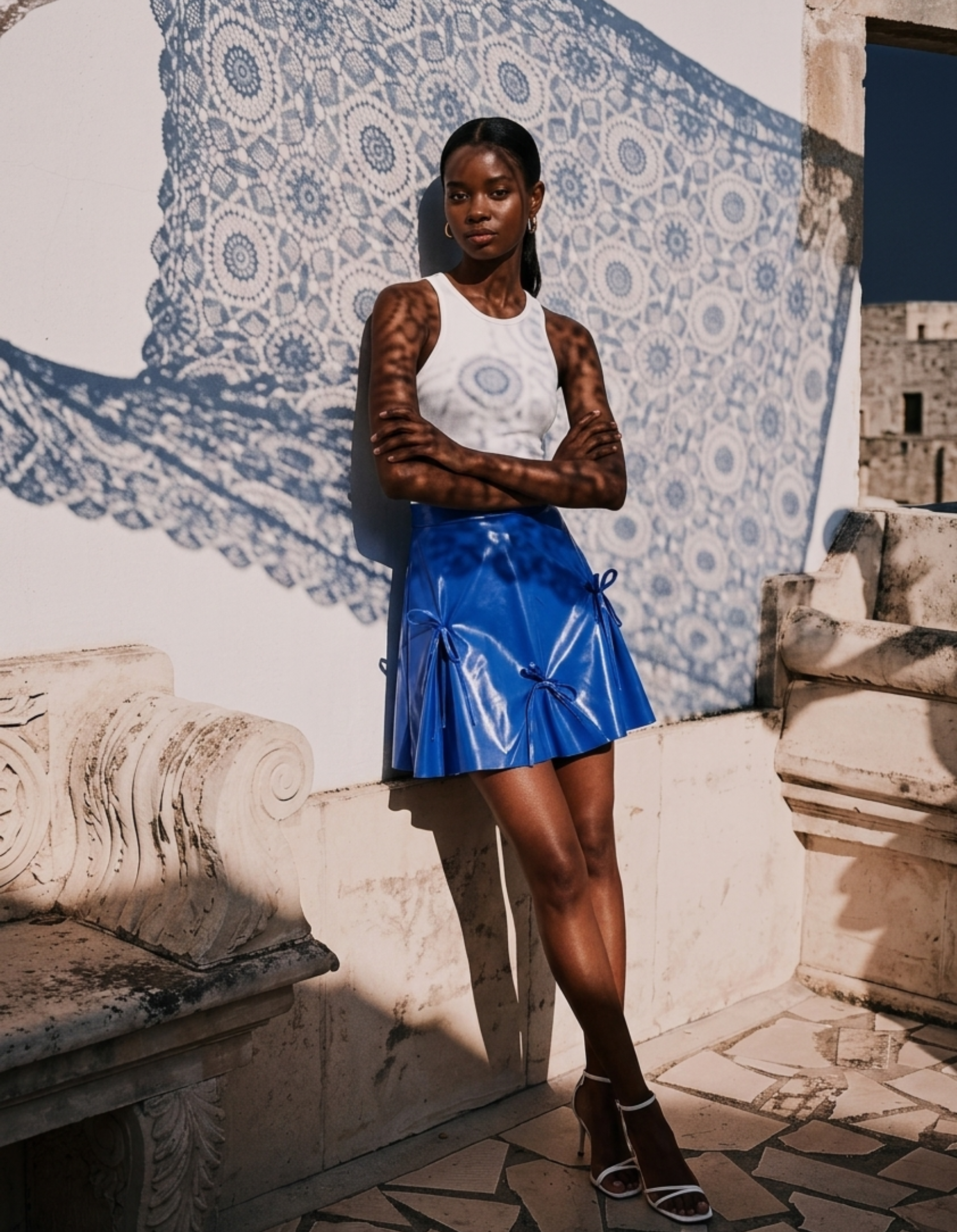 Woman in a white top and blue kingdom of daughters skirt standing against a decorative wall.