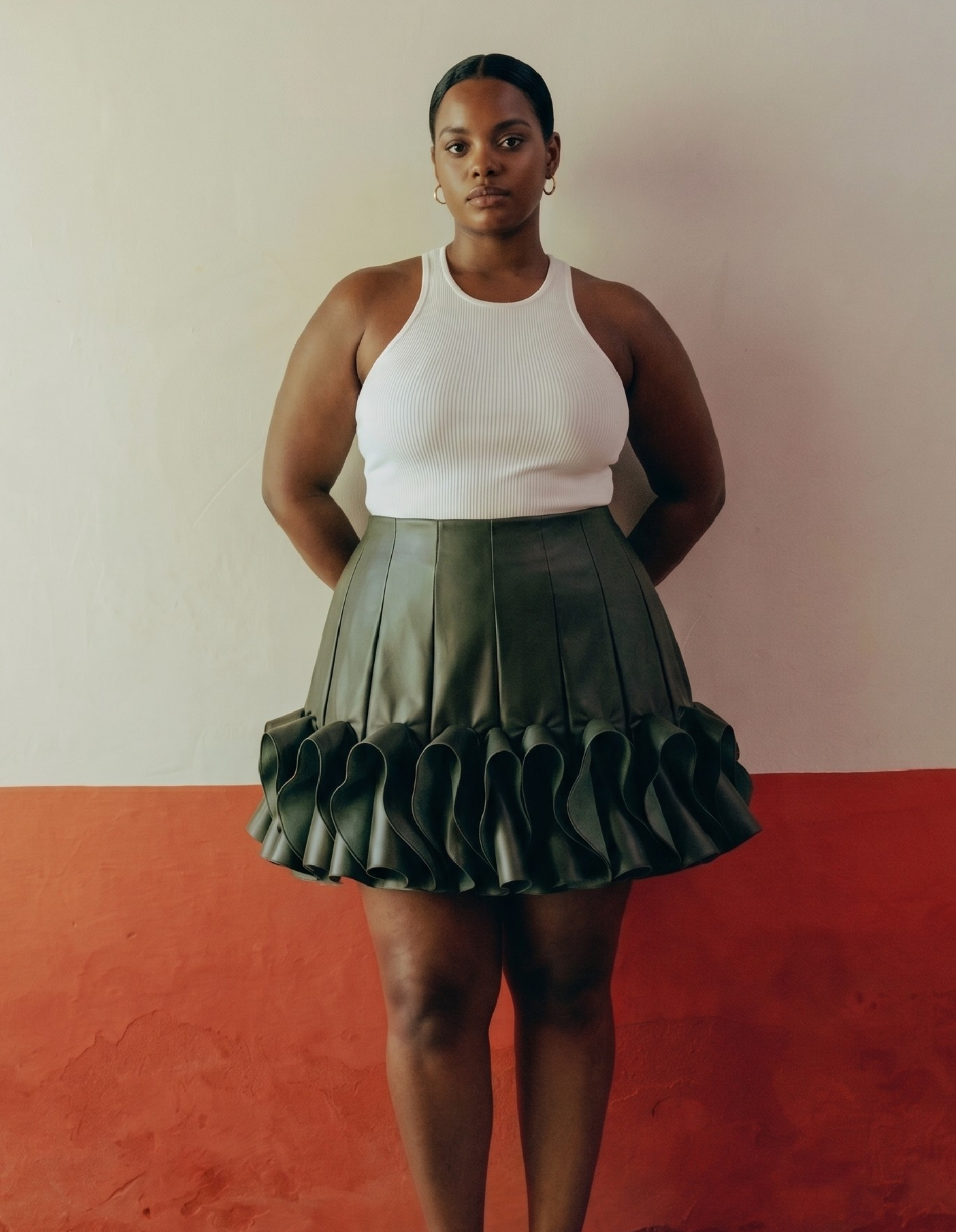 Person wearing a white tank top and green pleated kingdom of daughters skirt standing against a plain background.